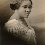 Black and white photographic bust length portrait of Madam C.J. Walker. Sitting sideways, her head is turned slightly right towards the viewer.